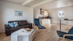 Ein modernes Wohnzimmer mit einem braunen Ledersofa, einem Glastisch und einem blauen Sessel. Im Hintergrund ist ein Küchenbereich mit weißen Schränken und einem Fenster erkennbar.