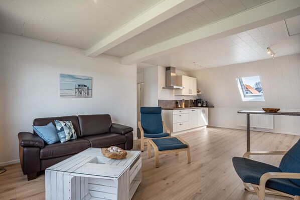 Un salon moderne avec un canapé en cuir marron, une table basse en verre et un fauteuil bleu. On aperçoit, au fond, un coin cuisine avec des meubles blancs et une fenêtre.