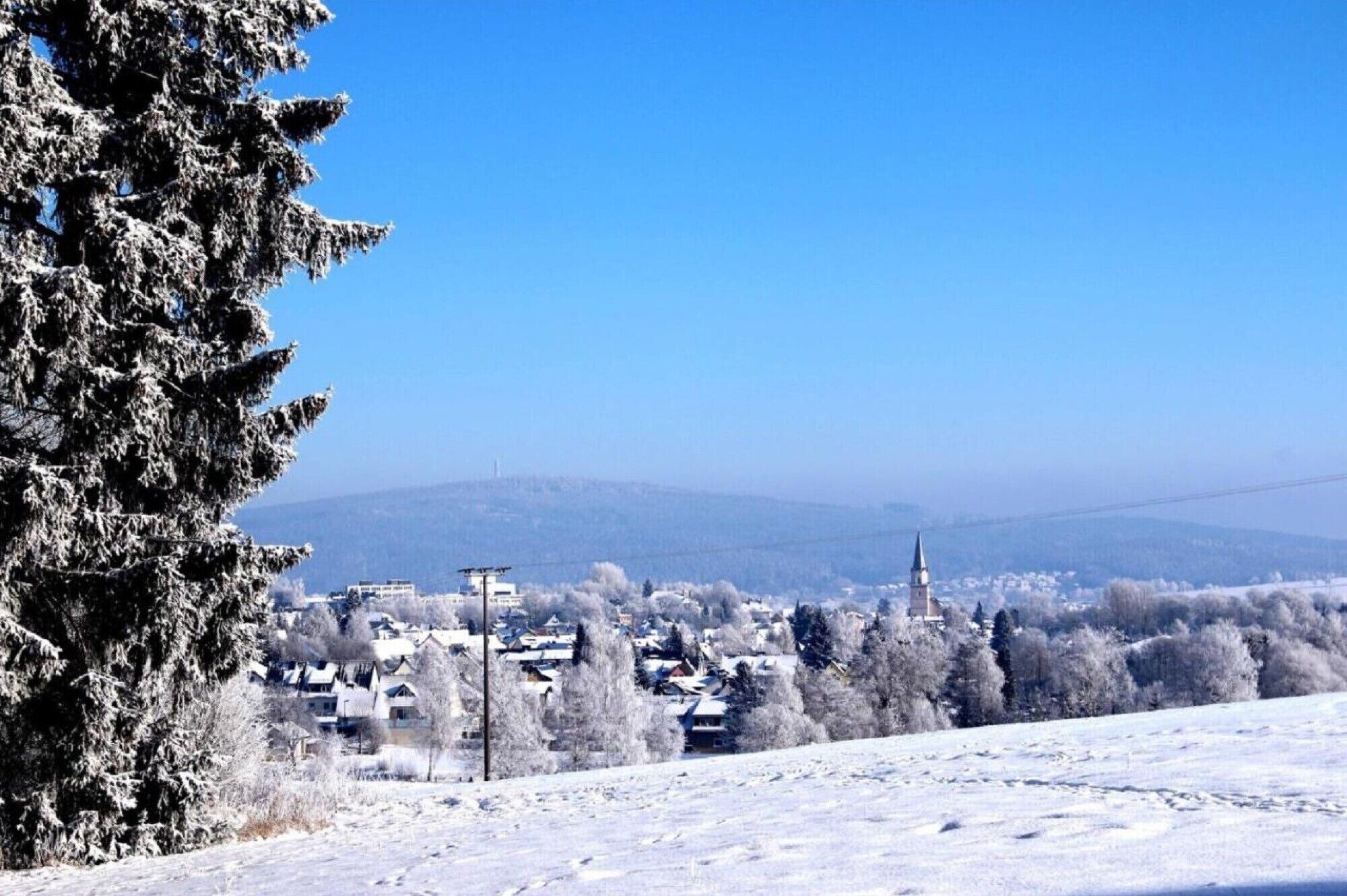 Schneebedeckte Landschaft mit einer Stadt in der Ferne, inklusive Häusern und einem Kirchturm.