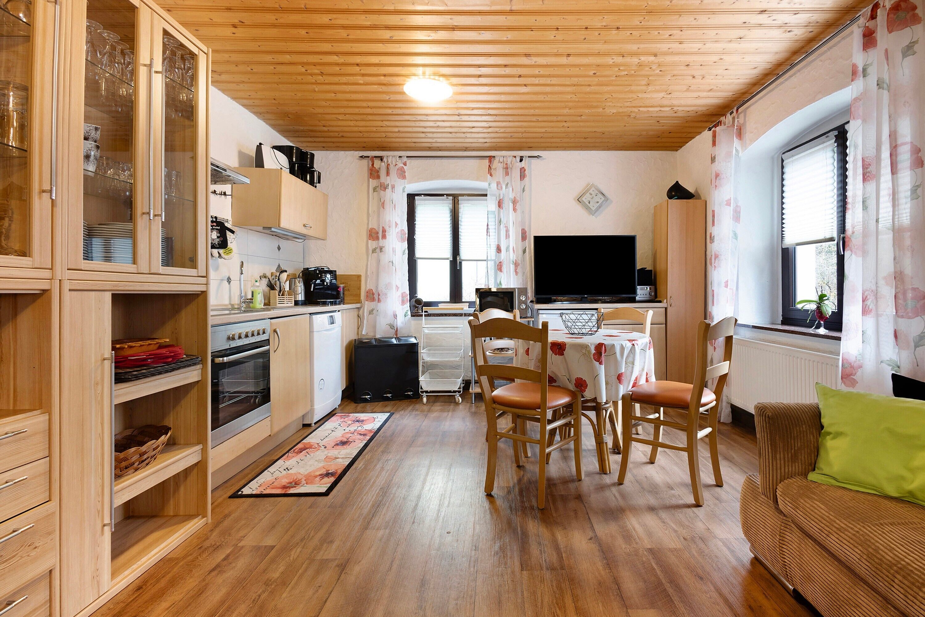 A compact kitchen with wooden cabinets, a dining area with chairs, a television, and a sofa with a green pillow.