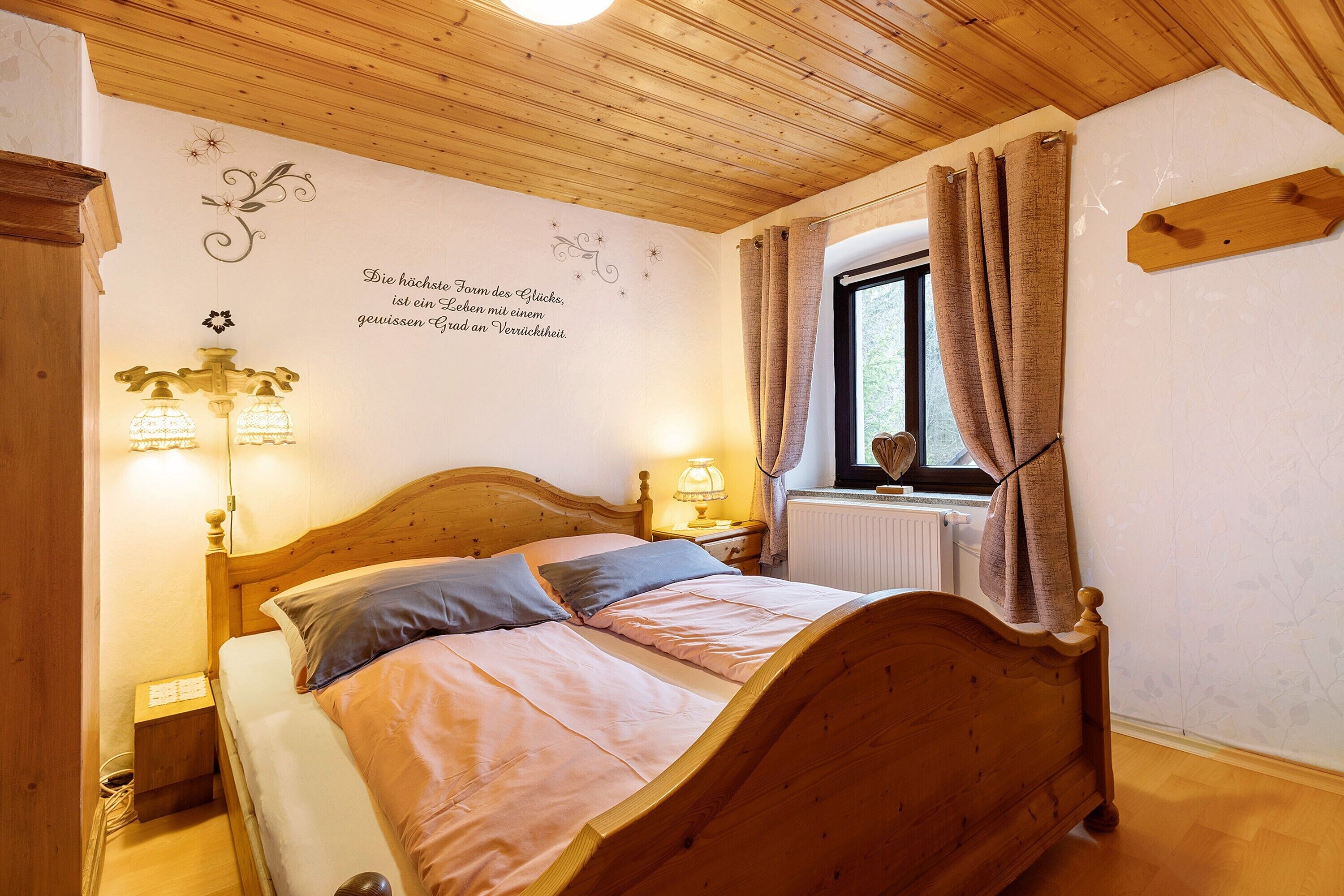 Ein Bett mit Holzrahmen, zwei Kissen, ein Nachttisch mit Lampe, ein Fenster mit Vorhängen und eine Wand mit dekorativem Spruch.