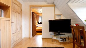 Een kamer met een televisie op een glazen voetstuk, houten meubels en een zichtbare deur naar een andere kamer.