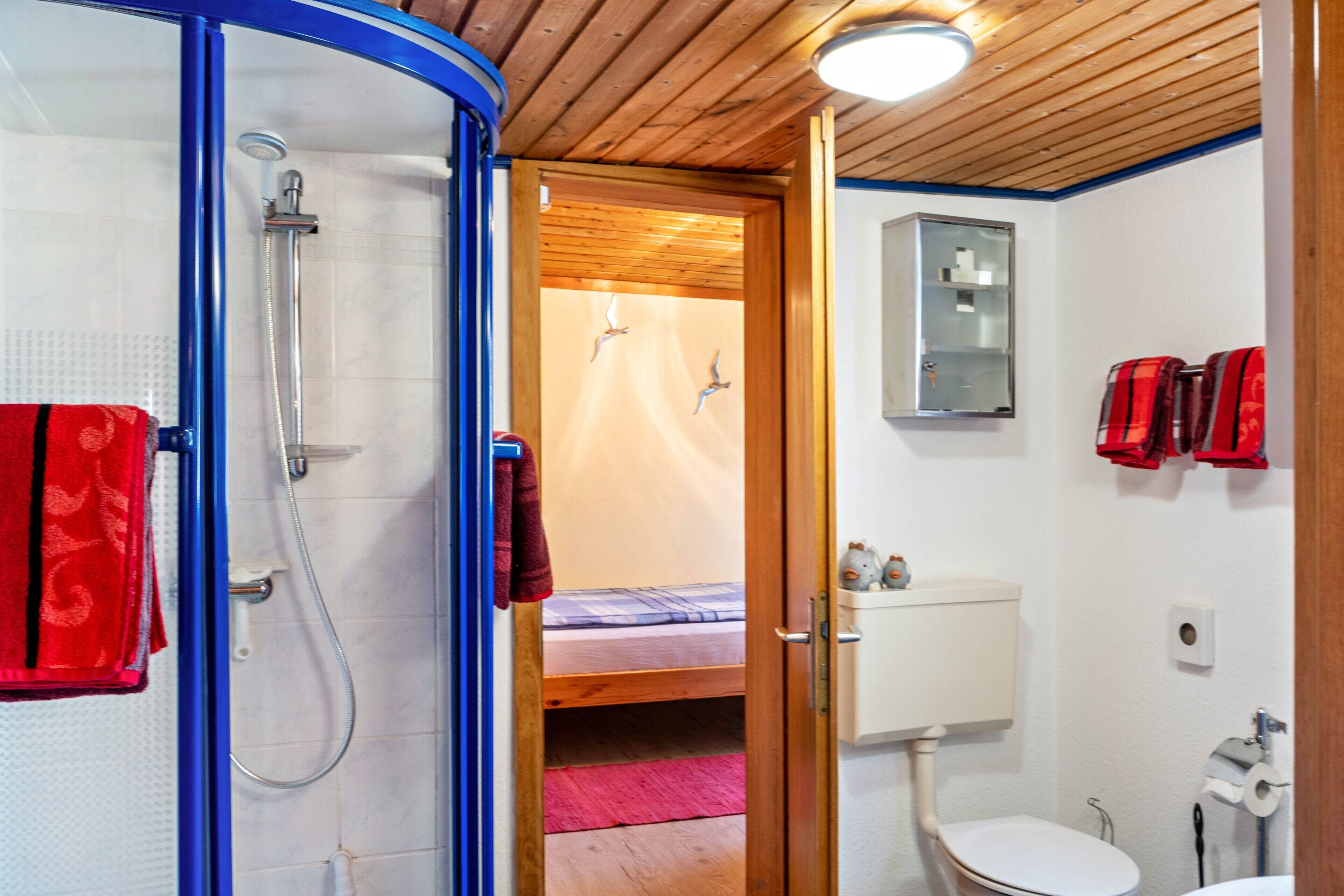 Een badkamer met douche, toilet en wastafel. In de aangrenzende kamer staat een bed.