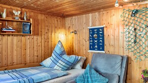 Interior de una cabaña de madera con una cama, una silla, un estante con adornos y una pared con un cartel de temática náutica.