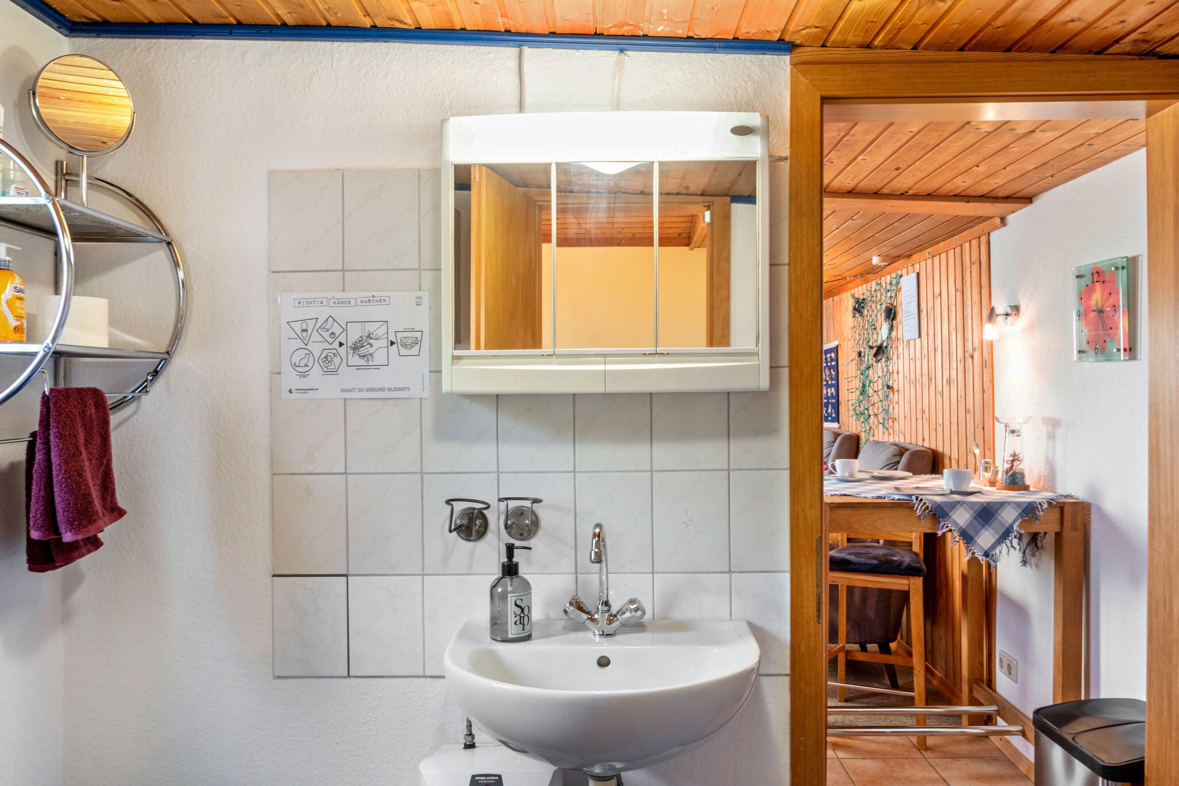 Ein Badezimmer mit Waschbecken, Spiegel und gefliesten Wänden. Eine Holzbalkendecke und ein Essbereich sind durch eine geöffnete Tür sichtbar.