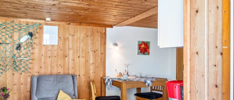 Una habitación con paneles de madera, una mesa de comedor, sillas, una cafetera y un sillón gris con un cojín amarillo.