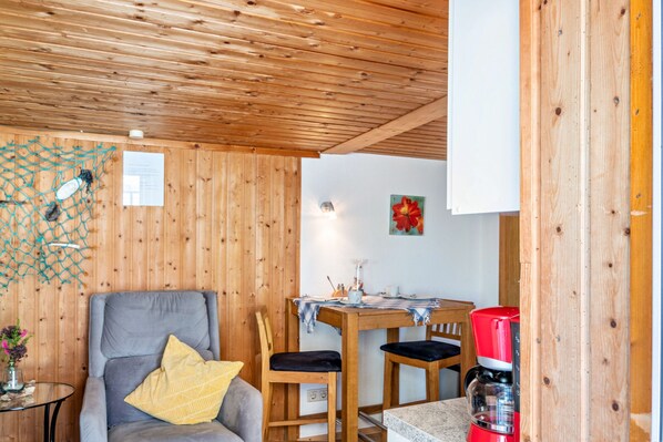 A wooden-paneled room with a dining table, chairs, a coffee maker, and a gray armchair with a yellow pillow.