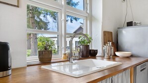 Una cocina con un lavabo blanco, una cafetera, una planta en maceta y una ventana con vista a árboles.