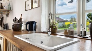 Una cocina con un lavamanos blanco grande, una cafetera y un ventanal con vistas a una casa y follaje.