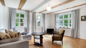 Una habitación con sofá, una mesa redonda, una silla, vigas de madera en el techo y ventanas grandes con cortinas.