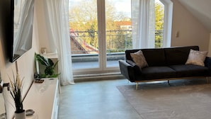A modern living room with a large window, a black sofa, a flat-screen TV, and a chandelier. - Apartment 'Maisonette Wohnung Frechen City' with Private Terrace, Wi-Fi and Air Conditioning (Frechen)