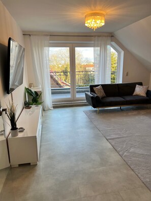 Ein modernes Wohnzimmer mit großem Fenster, einer schwarzen Couch, einem Flachbildfernseher und einem Chandelier.