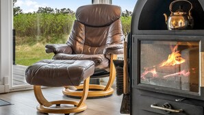 Ein modernes Wohnzimmer mit Ledersessel und Hocker, einem Kaminofen und einem großen Fenster mit Blick auf eine natürliche Landschaft.