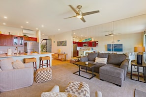 A modern living room with a grey sofa, ottoman, and coffee table. There is a kitchen area with red cabinets and a dining area with a table and chairs. - Oceanfront, updated, clean, modern. Great reviews! (Lahaina)