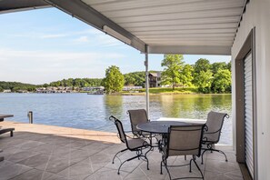 Eine überdachte Terrasse mit Tisch und Stühlen mit Blick auf einen See, im Hintergrund Häuser und Bäume.