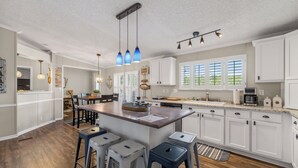 Una cocina moderna con armarios blancos, una isla central, taburetes de bar, lámparas colgantes y vistas al exterior a través de amplios ventanales.