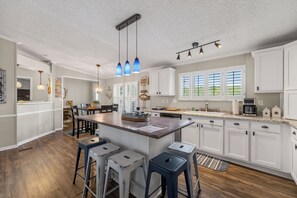 A modern kitchen with white cabinets, a central island, bar stools, pendant lights, and a view of the outdoors through large windows.