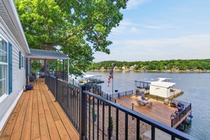 Una terraza de madera con mobiliario exterior, un espacio de estar cubierto y vista a un lago con barcos y un muelle.
