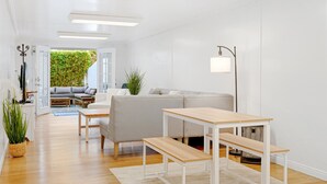 En moderne stue med sofa, sofabord, spisebord med bænke, gulvlampe og udsigt til en grøn have gennem en åben dør.