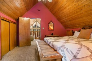 A bedroom with a sloped ceiling, a bed with a patterned bedspread, a wooden bench, a nightstand, and a mirror on the wall. - Memory Mtn Lodge by Velo (Angel Fire)