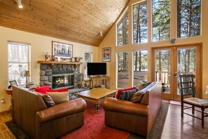 A cozy living room with a stone fireplace, leather sofas, and large windows offering a view of the forest. - Memory Mtn Lodge by Velo (Angel Fire)
