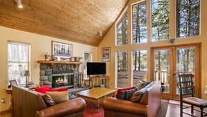 Una sala de estar acogedora con chimenea de piedra, sofás de cuero, una mesa de centro de madera y amplias ventanas con vistas al bosque circundante.
