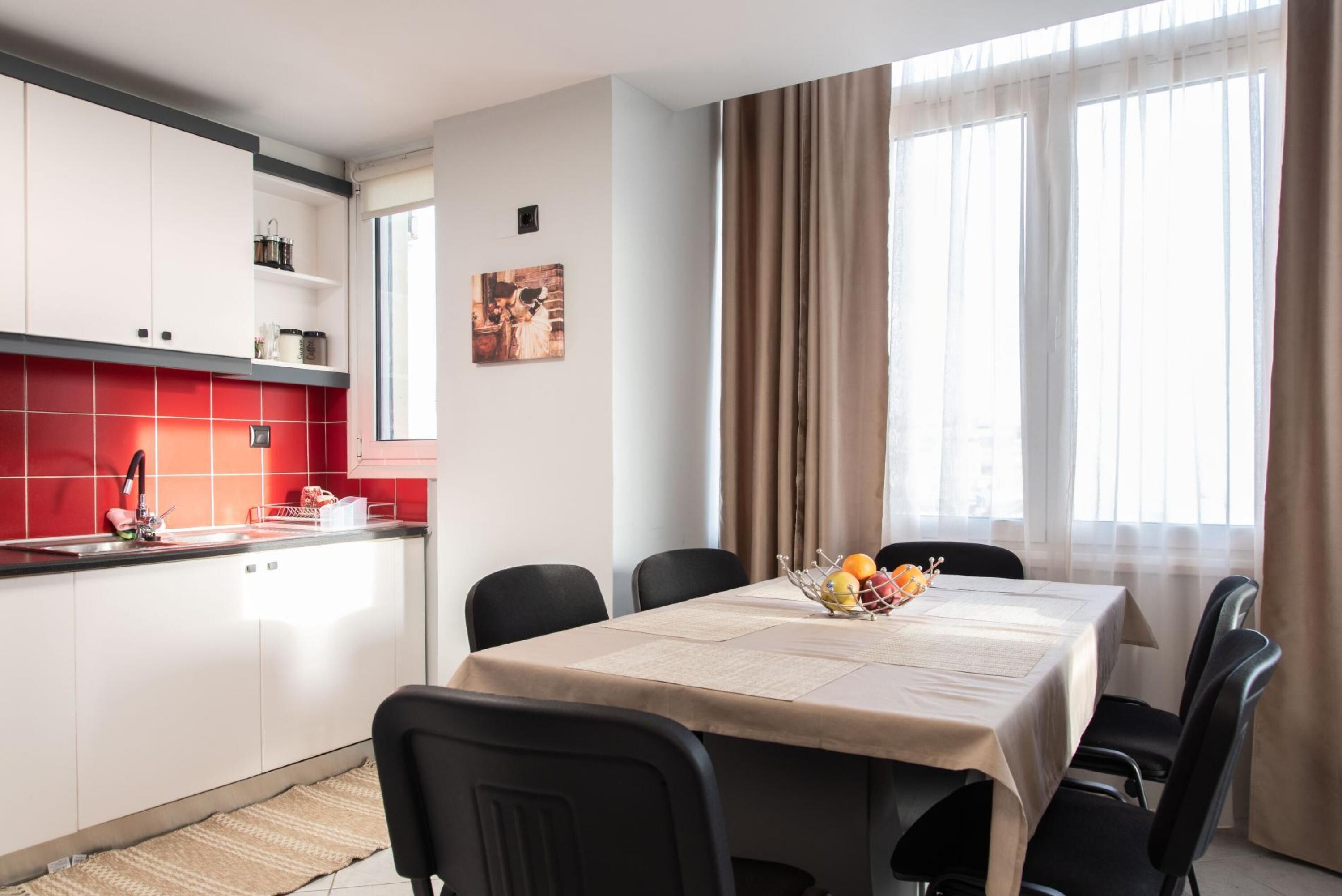 Una cocina moderna con armarios blancos y baldosas rojas, una mesa de comedor con mantel beige y un frutero, y un ventanal con cortinas.