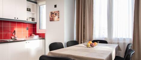 Una cocina moderna con armarios blancos y baldosas rojas, una mesa de comedor con mantel beige y un frutero, y un ventanal con cortinas.