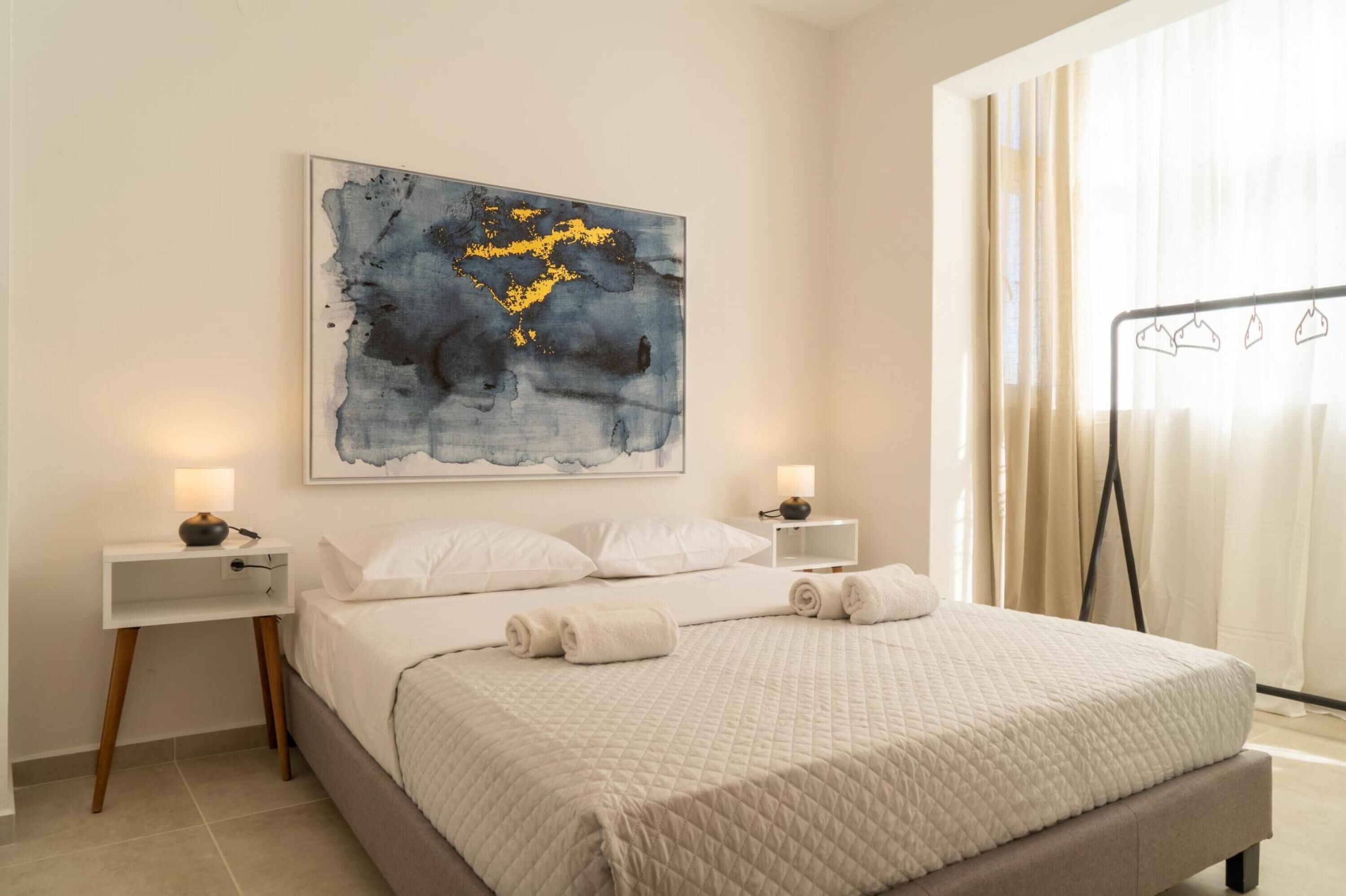Un lit bien fait, recouvert d’une couette blanche matelassée, deux tables de chevet avec des lampes et un grand tableau abstrait accroché au mur.
