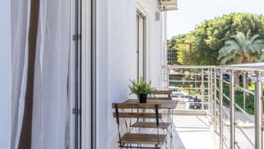 A balcony with a table and chairs, a potted plant, and a view of a street with cars and trees. - Pananos Sq House I (Heraklion)