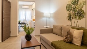 A living room with a sofa, a coffee table, and a plant. There is a door and a window with curtains. - Pananos Sq House II (Heraklion)