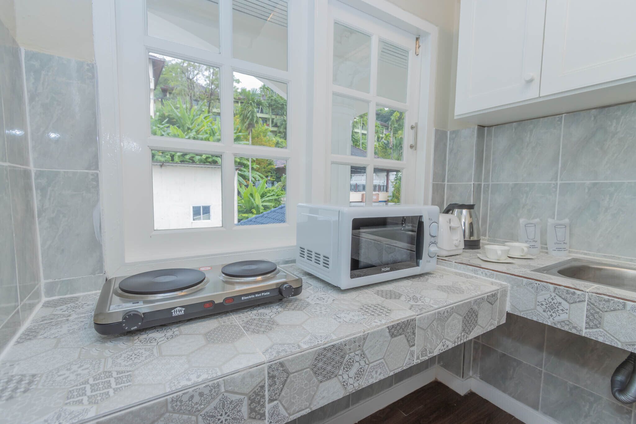 A kitchen with a microwave, electric hot plate, and a window with a view of greenery.