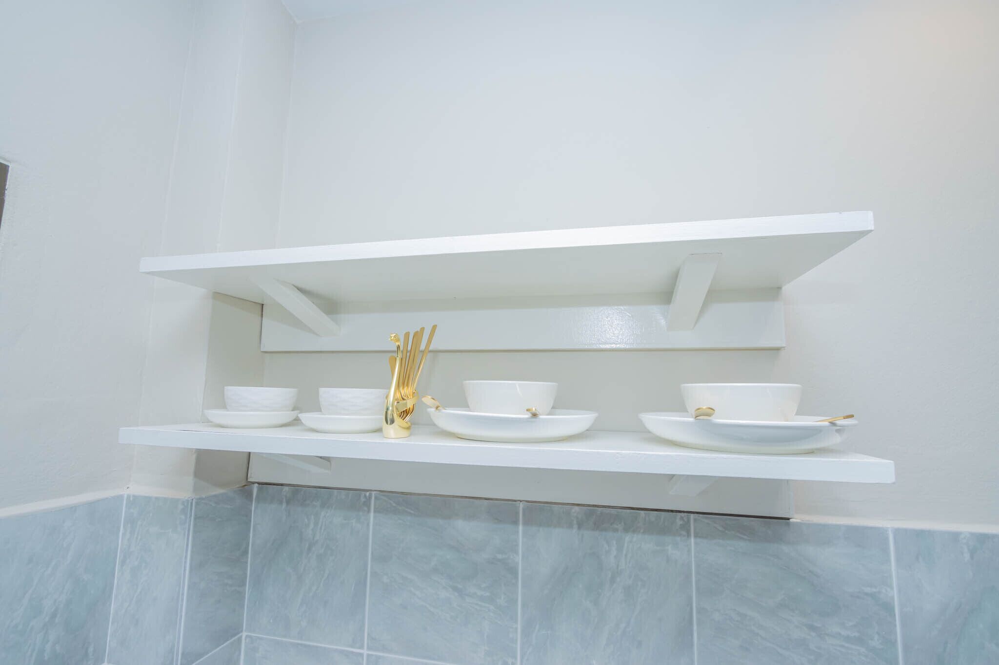 A white shelf with white bowls and a gold utensil holder in a bathroom with light blue tiles.