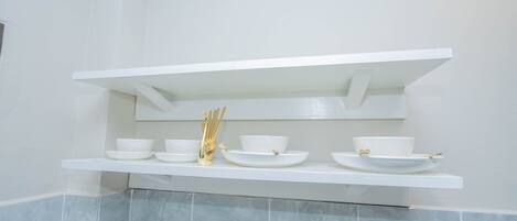 A white shelf with white bowls and a gold utensil holder in a bathroom with light blue tiles.