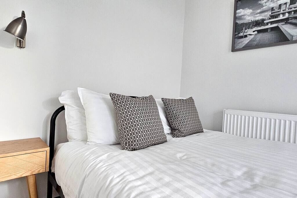 A neatly made bed with white and patterned pillows, a bedside table, a wall-mounted lamp, and a framed picture on the wall.