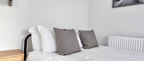 A neatly made bed with white and patterned pillows, a bedside table, a wall-mounted lamp, and a framed picture on the wall.