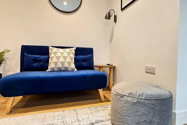 Ein blaues Sofa mit gemusterten Kissen, ein kleiner Holztisch, ein grauer Hocker und ein runder Spiegel an der Wand.