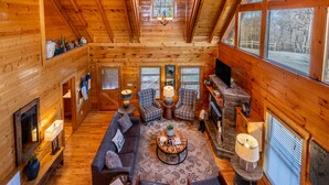 A cozy log cabin interior with a fireplace, wooden walls, and large windows offering a view of the outdoors. - Misty Mountain by AvantStay | Hot Tub, Mountain Views, Theater, Pool Table (Sevierville)