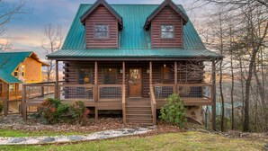 A log cabin with a green roof, a porch with a railing, and a stone foundation. - Misty Mountain by AvantStay | Hot Tub, Mountain Views, Theater, Pool Table (Sevierville)