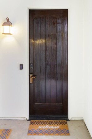 Una puerta de madera oscura con un picaporte de latón, una lámpara en la pared a la izquierda y un felpudo en la entrada.