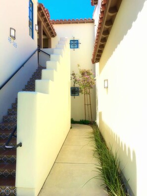 Un étroit sentier mène à un escalier dont le sol est recouvert de carreaux et qui est bordé d’un mur blanc percé d’une porte.