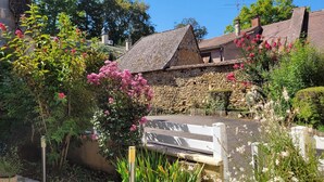 A stone building with a sloped roof, surrounded by lush greenery and flowering plants. - Charmant Gîte 3 Cosy, Climatisé, Environnement Bucolique, en Périgord Noir (Sagelat)