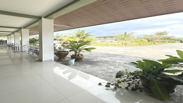 A covered outdoor area with tiled flooring, potted plants, and a view of palm trees and a building in the distance. - OYO 75467 Baan Siripornchai 2 (Sakon Nakhon)