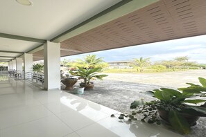 A covered outdoor area with tiled flooring, potted plants, and a view of palm trees and a building in the distance. - OYO 75467 Baan Siripornchai 2 (Sakon Nakhon)