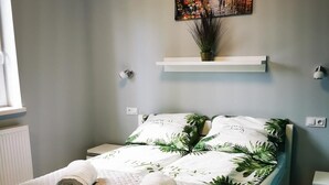 Una cama bien hecha con una cubierta acolchada, dos toallas enrolladas y una funda de almohada con motivos tropicales. Encima de la cama, un cuadro de una calle de la ciudad.