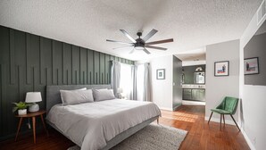 A bedroom with a bed, bedside tables, a ceiling fan, and a view into the kitchen.