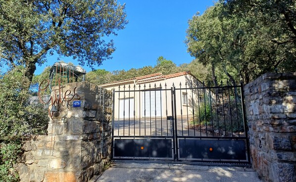 Una entrada con reja a un edificio, con un muro de piedra y portón de metal, rodeada de árboles.