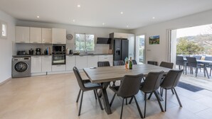 Une cuisine moderne avec des meubles blancs, un coin repas doté d’une grande table et de chaises, et une vue sur l’extérieur grâce à une porte-fenêtre coulissante.