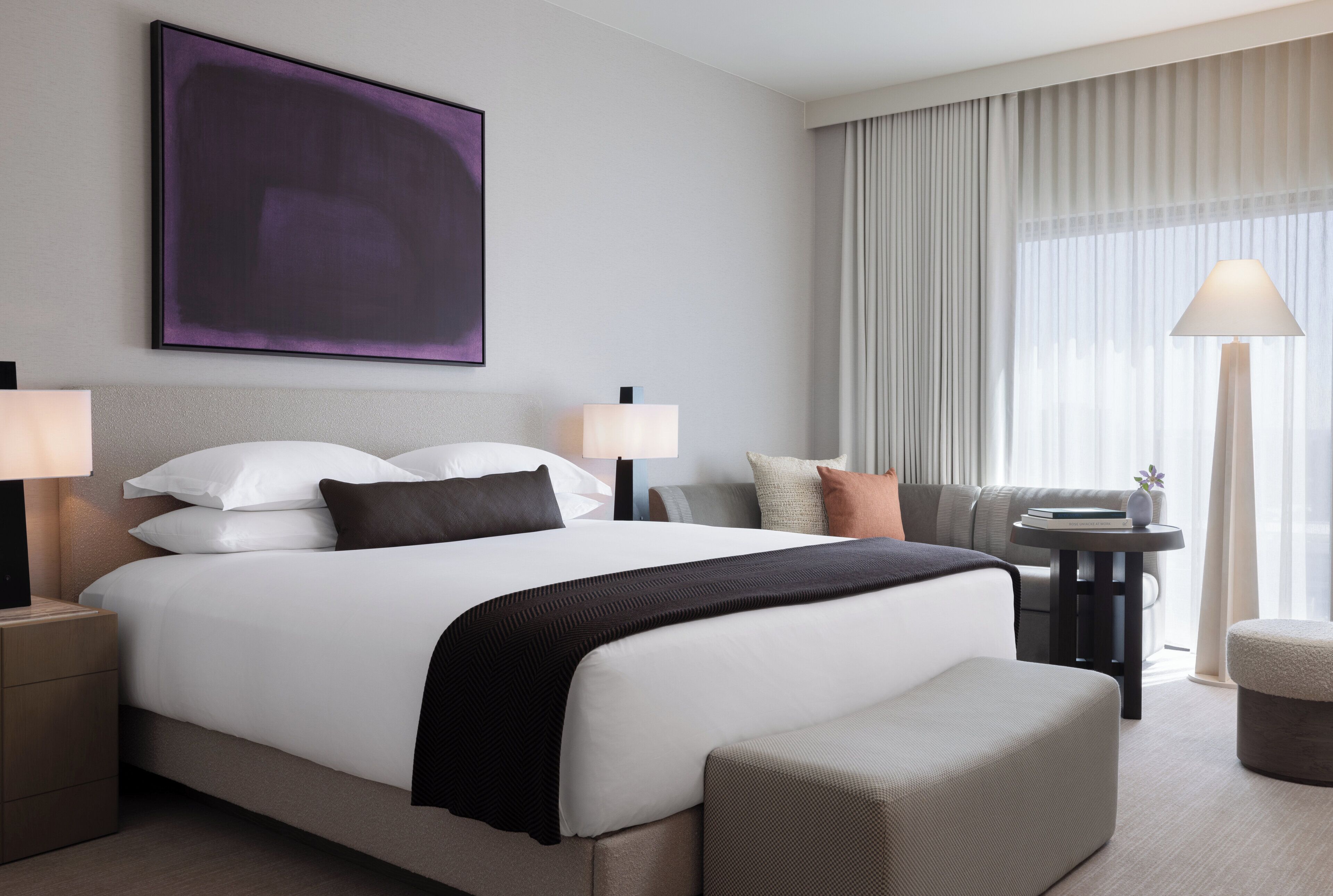 Ett modernt hotellrum med en stor säng, två sängbord, en soffa, ett litet bord med en lampa och ett stort fönster med gardiner.