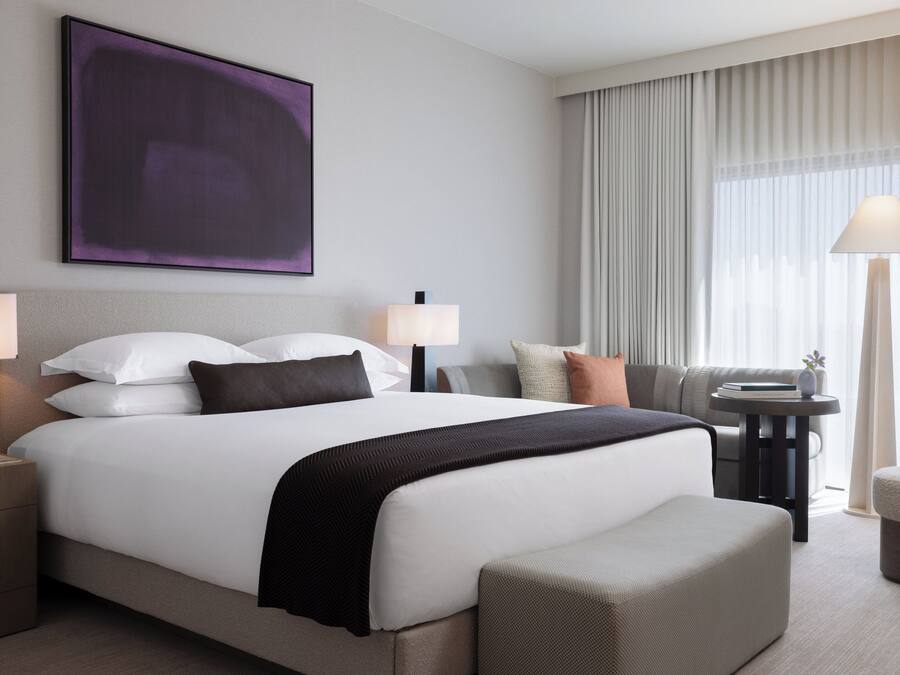 Ett modernt hotellrum med en stor säng, två sängbord, en soffa, ett litet bord med en lampa och ett stort fönster med gardiner.